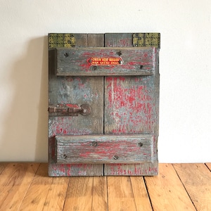 May include: A rustic wooden wall decor piece with a red, white and blue colour scheme. The piece is made of reclaimed wood and has a distressed finish. It features a metal latch and a label that reads "Crown Rom Brand Man Akcese Ana".