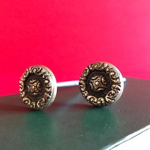 Könnte beinhalten: Zwei goldfarbene Metall-Schubladengriffe mit verzierten Designs. Die Griffe sind kreisförmig mit einer erhabenen Mitte und einem dekorativen Rand.