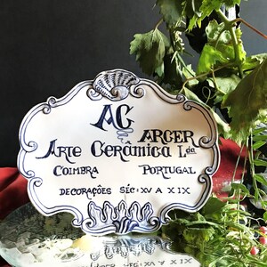 May include: A white ceramic plaque with blue decorative trim and text. The plaque reads "AG ARCER Arte Cerâmica Lda COIMBRA PORTUGAL DECORAÇÕES SÉC: XV A XIX". The plaque has a seashell design at the top.
