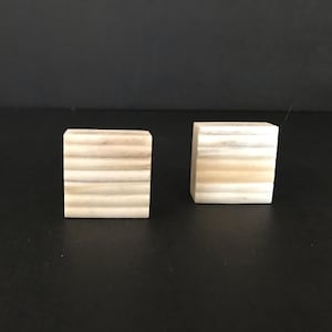 May include: Two small, square, wooden blocks with a layered, striped pattern. The blocks are a light beige colour with darker beige stripes.