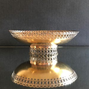Op de afbeelding: Een goudkleurige metalen schaal op voet met een brede, ondiepe kom en een decoratieve, opengewerkte rand. De schaal wordt ondersteund door een cilindrische voet die eveneens is voorzien van een bijpassend, ingewikkeld uitgesneden patroon.