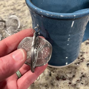 May include: A silver mesh tea infuser with a hinged design, held near a blue ceramic mug. The infuser has a small hook for hanging. The mug has a wide rim and a curved handle.