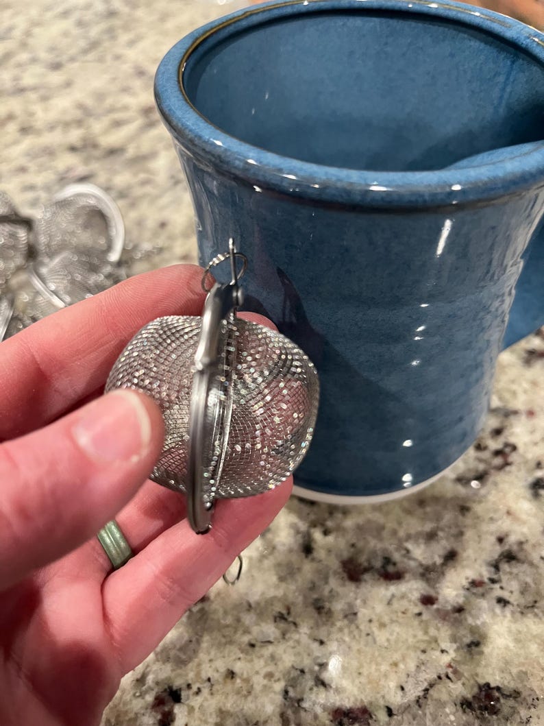 May include: A silver mesh tea infuser with a hinged design, held near a blue ceramic mug. The infuser has a small hook for hanging. The mug has a wide rim and a curved handle.