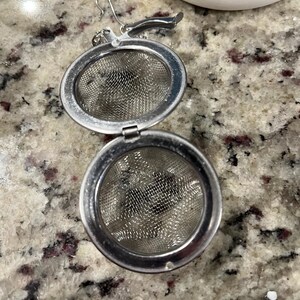 May include: A stainless steel tea infuser with a hinged, round design. The infuser is open, revealing a fine mesh interior. A small handle and hook are attached for easy use. The background shows a granite countertop and a blue and white ceramic object.