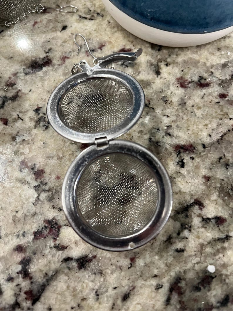 May include: A stainless steel tea infuser with a hinged, round design. The infuser is open, revealing a fine mesh interior. A small handle and hook are attached for easy use. The background shows a granite countertop and a blue and white ceramic object.