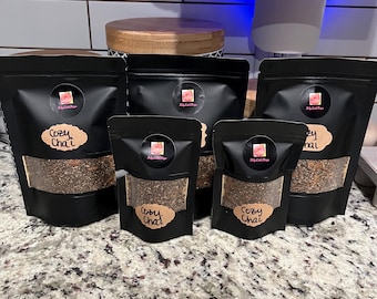 May include: Several black pouches of Cozy Chai tea are arranged on a countertop. Each pouch has a clear window showing the tea blend and a sticker with a logo. The words "Cozy Chai" are written on a brown label.