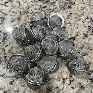 May include: A collection of stainless steel tea infusers. Each infuser is a small, spherical mesh basket with a hinged opening and a chain for easy removal from a cup or teapot. The infusers are scattered on a speckled surface.