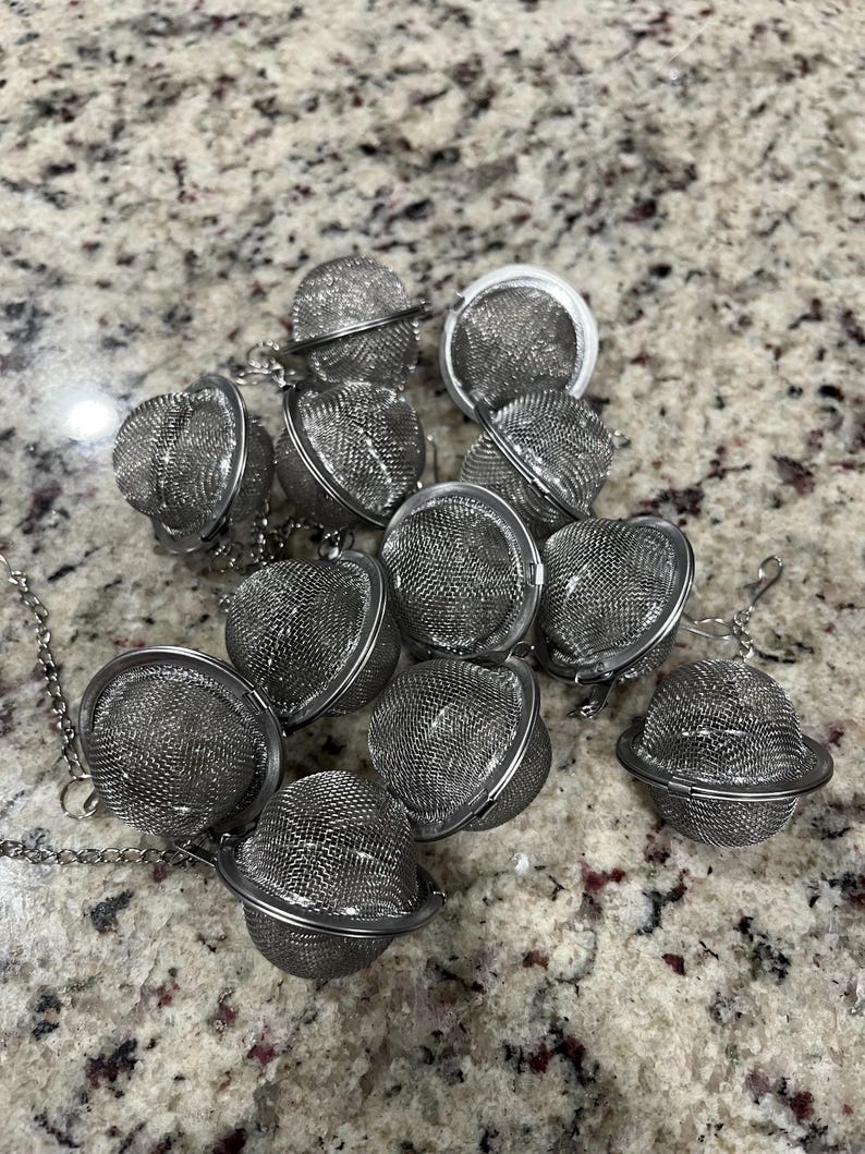 May include: A collection of stainless steel tea infusers. Each infuser is a small, spherical mesh basket with a hinged opening and a chain for easy removal from a cup or teapot. The infusers are scattered on a speckled surface.