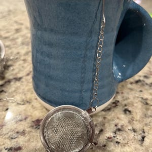 May include: A teal ceramic mug with a white rim, paired with a stainless steel tea infuser. The infuser is a round, mesh basket attached to a chain. The mug has a large handle and sits on a speckled surface.