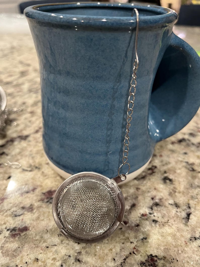 May include: A teal ceramic mug with a white rim, paired with a stainless steel tea infuser. The infuser is a round, mesh basket attached to a chain. The mug has a large handle and sits on a speckled surface.