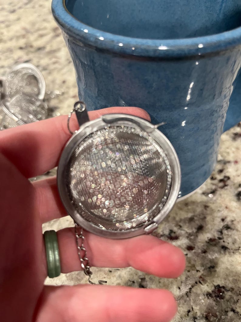May include: A stainless steel tea infuser with a fine mesh basket and a chain. The infuser is held near a blue ceramic mug. The infuser is designed to hold loose leaf tea for steeping.