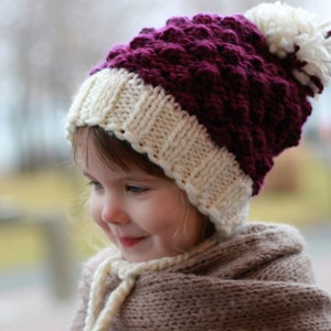 May include: A child wearing a white and burgundy knitted hat with a white pom pom. The hat has a textured design and is folded down over the child's ears.