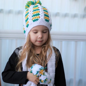 May include: A young girl wearing a white crocheted hat with green and yellow stripes and a matching scarf. The hat has a pom-pom on top.