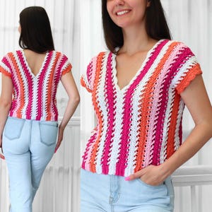 May include: A short-sleeved crochet top featuring vertical stripes in shades of bright pink, orange, white, and light pink. The garment has a V-neckline, a scalloped edge along the sleeves, and a textured, open-stitch pattern throughout the body.