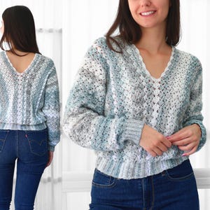 Puede incluir: Un suéter con cuello en V en tonos de azul, blanco y gris. La parte superior de manga larga tiene un diseño texturizado de tejido abierto y un dobladillo y puños acanalados. El suéter se combina con unos vaqueros azules.