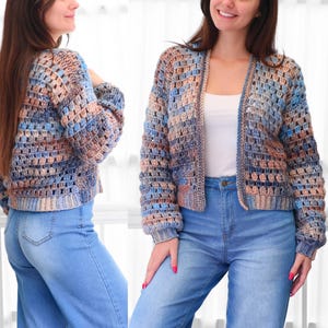 May include: A multi-colored, open-front crochet cardigan in shades of blue, tan, and brown. The handmade sweater features a loose, open weave design with long sleeves and a relaxed fit. The cardigan is paired with blue jeans.