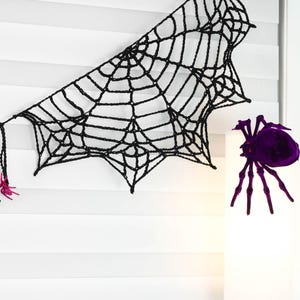 May include: A black crocheted spider web decoration featuring a series of interconnected yarn strands. The web is accented with small, bright pink glittery spiders attached to the edges and a large, dark purple spider positioned nearby.