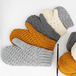 May include: A set of five crocheted mittens in various colors, including gray, beige, and mustard yellow. The mittens are arranged in a semi-circle, with the gray mitten in the foreground. There are also two balls of yarn in the background, one mustard yellow and one white.