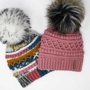 May include: Two crocheted hats with faux fur pom poms. One hat is pink with a black and white pom pom and the other is multicolored with a white pom pom.
