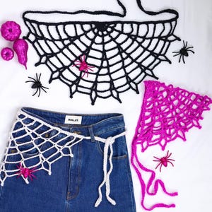Puede incluir: Un top de ganchillo negro con diseño de telaraña, un cinturón de ganchillo blanco y un pañuelo de ganchillo rosa brillante. Decorado con pequeñas arañas de plástico negras, arañas rosas brillantes y calabazas rosas con purpurina. El cinturón se coloca sobre unos vaqueros azules con la etiqueta Rolla's.