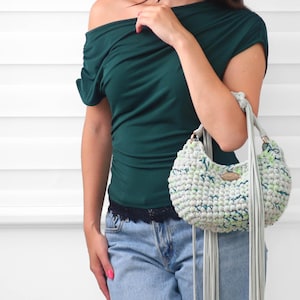 May include: A dark green off-the-shoulder top with ruched detailing, light blue denim jeans with a black lace trim, and a light gray crocheted handbag featuring long fringe accents, a rounded shape, and a small oval-shaped metallic brand tag.