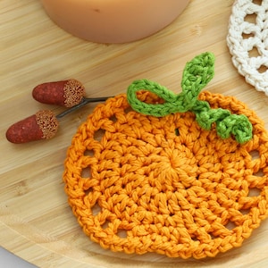 May include: A round orange crochet coaster shaped like a pumpkin with a green crocheted stem and leaf detail. Two small brown acorns with textured caps are positioned to the left of the pumpkin coaster, which features an openwork stitch pattern.