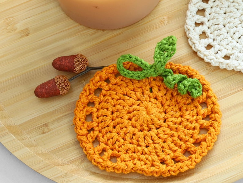 May include: A round orange crochet coaster shaped like a pumpkin with a green crocheted stem and leaf detail. Two small brown acorns with textured caps are positioned to the left of the pumpkin coaster, which features an openwork stitch pattern.