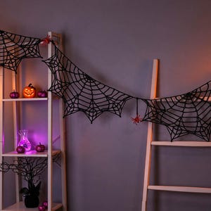 May include: A black crocheted spider web garland featuring intricate web patterns and small red spider accents. The garland is draped across a white wooden shelving unit and a white ladder, creating a decorative display with a dark, textured aesthetic.