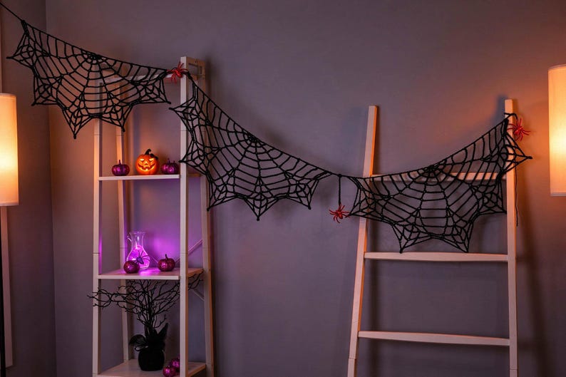 May include: A black crocheted spider web garland featuring intricate web patterns and small red spider accents. The garland is draped across a white wooden shelving unit and a white ladder, creating a decorative display with a dark, textured aesthetic.