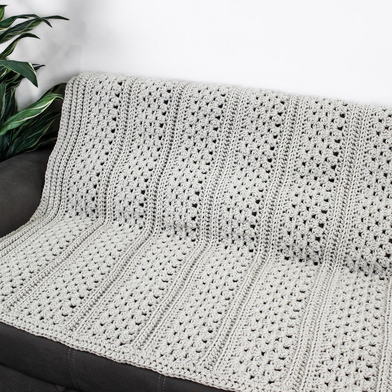 May include: A gray crocheted throw blanket with a textured pattern. The blanket is draped over a gray couch.