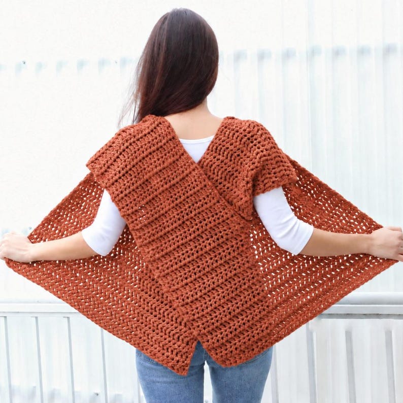 May include: A rust-colored, hand-crocheted wrap vest featuring a textured, ribbed stitch pattern. The garment has a wide, overlapping front design that creates a triangular silhouette, worn over a white short-sleeved shirt and blue denim jeans.