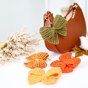 May include: A brown crescent-shaped handbag with a gold-tone metal clasp, adorned with a textured olive green crochet bow keychain. Three additional crochet bows in shades of mustard yellow, burnt orange, and bright orange lie nearby.