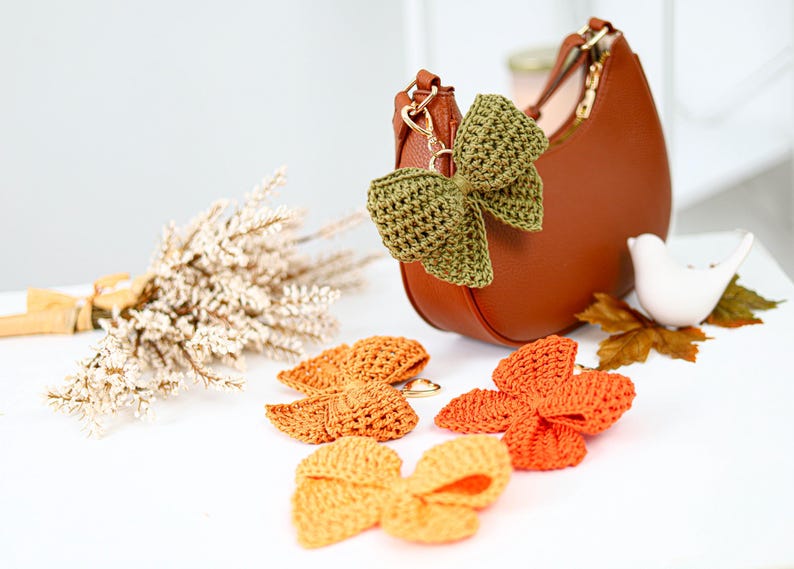 May include: A brown crescent-shaped handbag with a gold-tone metal clasp, adorned with a textured olive green crochet bow keychain. Three additional crochet bows in shades of mustard yellow, burnt orange, and bright orange lie nearby.