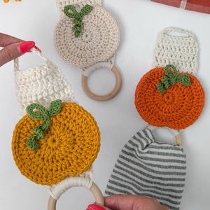 May include: Three crochet towel holders featuring round pumpkin motifs in cream, orange, and yellow. Each piece includes a wooden ring, a crocheted hanging loop, and a small green vine detail. One holder is attached to a gray and white striped towel.