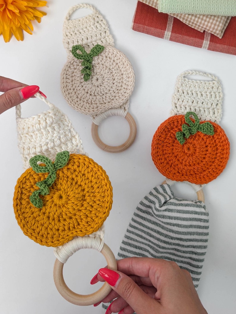 May include: Three crochet towel holders featuring round pumpkin motifs in cream, orange, and yellow. Each piece includes a wooden ring, a crocheted hanging loop, and a small green vine detail. One holder is attached to a gray and white striped towel.