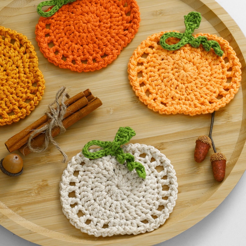 May include: Three crocheted pumpkin-shaped coasters in orange, light orange, and cream, each featuring a green stem. The set includes a bundle of three cinnamon sticks tied with twine, a single brown acorn, and two small acorns on a stem.