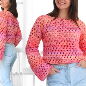 May include: A cropped, long-sleeved crochet sweater in a gradient of pink, orange, and purple hues. The open weave design is visible, and the sweater has a round neckline and flared sleeves. The model is wearing light blue jeans.
