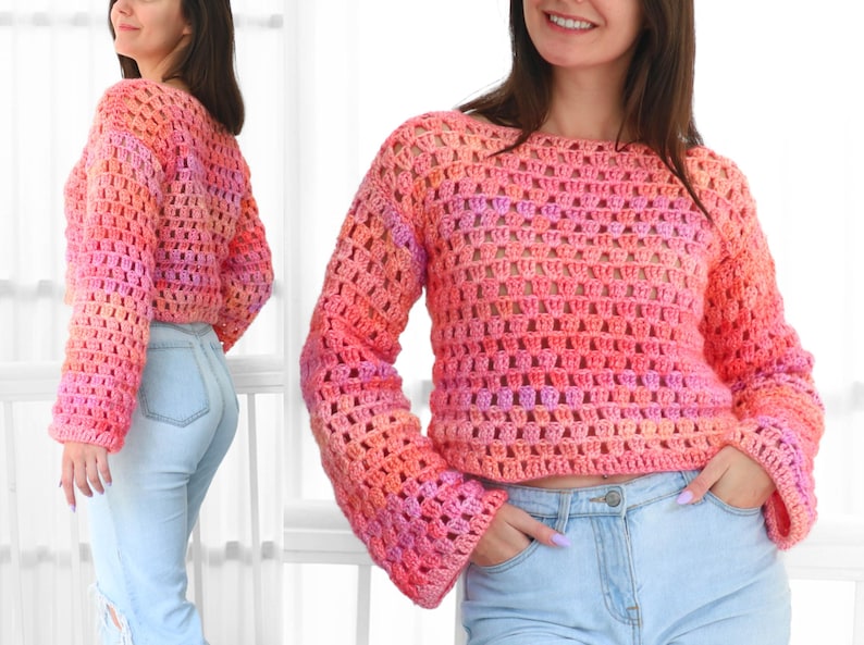 May include: A cropped, long-sleeved crochet sweater in a gradient of pink, orange, and purple hues. The open weave design is visible, and the sweater has a round neckline and flared sleeves. The model is wearing light blue jeans.