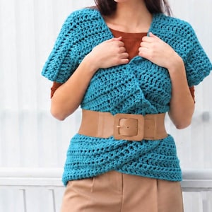 May include: A teal blue crochet top with short sleeves and a textured knit pattern, featuring a wide tan faux leather belt with a large square buckle worn around the waist over a solid brown short-sleeved shirt and tan trousers.