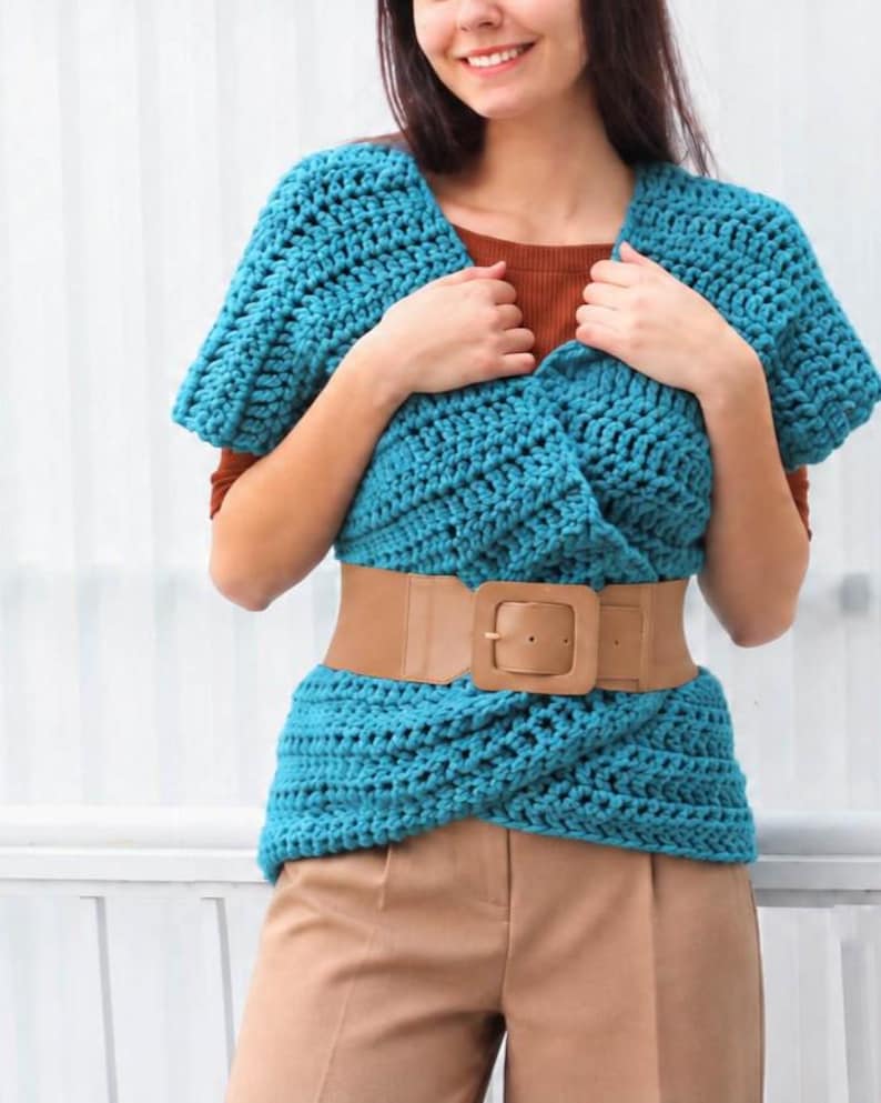 May include: A teal blue crochet top with short sleeves and a textured knit pattern, featuring a wide tan faux leather belt with a large square buckle worn around the waist over a solid brown short-sleeved shirt and tan trousers.