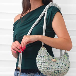 May include: A crescent-shaped crochet handbag crafted from light gray yarn with flecks of green and dark blue. The bag features a long, light gray strap with metallic gold-toned end caps and a small oval wooden tag labeled handmade on the front.