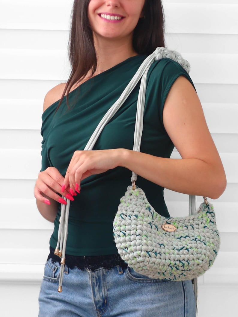 May include: A crescent-shaped crochet handbag crafted from light gray yarn with flecks of green and dark blue. The bag features a long, light gray strap with metallic gold-toned end caps and a small oval wooden tag labeled handmade on the front.