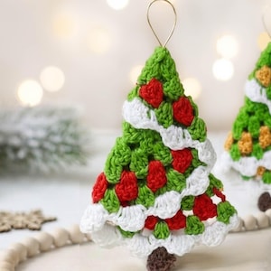 May include: Crocheted Christmas tree ornament with green, white, and red details. The tree features a green body with white and red accents, and a brown base. A gold loop is attached for hanging. Festive holiday decor.