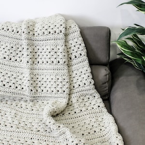 May include: A white crocheted throw blanket draped over a gray couch. The blanket has a textured, open-stitch pattern.