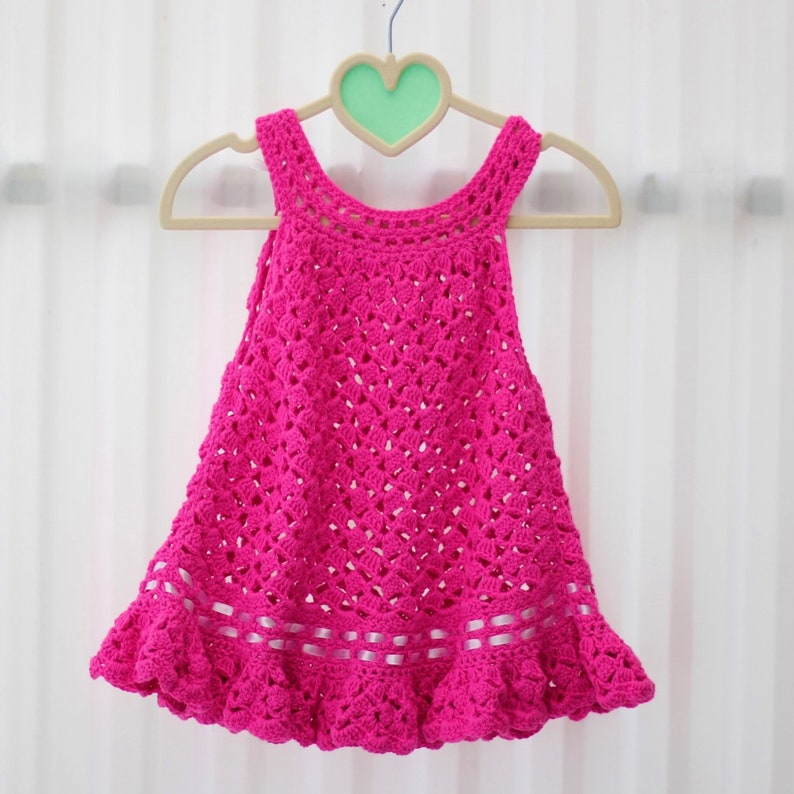 May include: A pink crocheted dress with a ruffled hem and white ribbon trim. The dress is hanging on a white hanger with a green heart.