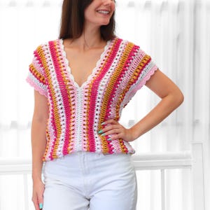 Pu&ograve; includere: Un top all'uncinetto colorato con scollo a V. Il top presenta un motivo a righe nei toni del rosa, bianco e oro. Le maniche e lo scollo sono rifiniti con un delicato bordo all'uncinetto rosa. Il top &egrave; abbinato a pantaloni bianchi.