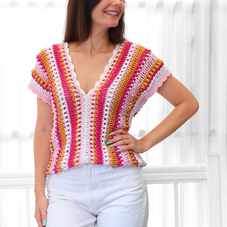 Pu&ograve; includere: Un top all'uncinetto colorato con scollo a V. Il top presenta un motivo a righe nei toni del rosa, bianco e oro. Le maniche e lo scollo sono rifiniti con un delicato bordo all'uncinetto rosa. Il top &egrave; abbinato a pantaloni bianchi.