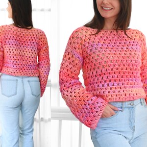 May include: A cropped, open-weave crochet sweater in shades of pink, orange, and purple. The sweater has long sleeves and a boat neck. The model is wearing light blue jeans. The sweater's texture and color gradient are prominent.