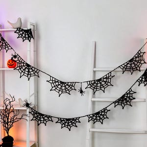 May include: A black crocheted spider web garland featuring multiple web segments connected by a thin string, with small black spider figures attached to the webs. The garland is draped across a white ladder-style shelf and a white wall.