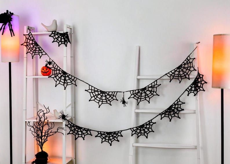 May include: A black crocheted spider web garland featuring multiple web segments connected by a thin string, with small black spider figures attached to the webs. The garland is draped across a white ladder-style shelf and a white wall.